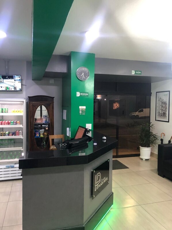 Un mostrador de recepción con un reloj, un letrero que indica 'Saída' (Salida) y un letrero que dice 'Recepção' (Recepción). Hay una planta en maceta y un cuadro en la pared.