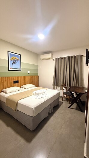 Habitación de hotel con cama, escritorio, silla, unidad de aire acondicionado y un cuadro en la pared.
