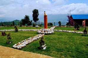 A garden with a central pillar, a fenced area, and a building with a blue roof. - Zambala Halesi Resort (Mahadevsthan)