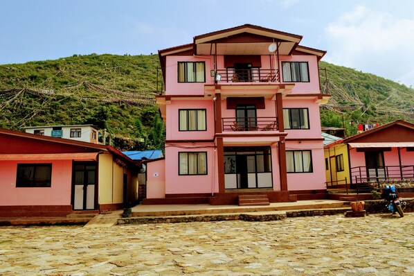 Un immeuble rose à plusieurs étages, doté de balcons, une cour pavée de pierres et un paysage montagneux en arrière-plan.