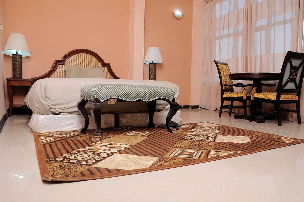 A hotel room with a bed, two bedside lamps, a table with chairs, and a patterned rug.
