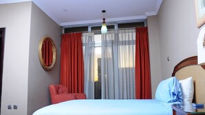 A bedroom with a bed, a chair, a mirror, and a window with curtains. - Holly Wood Hotel (Addis Ababa)