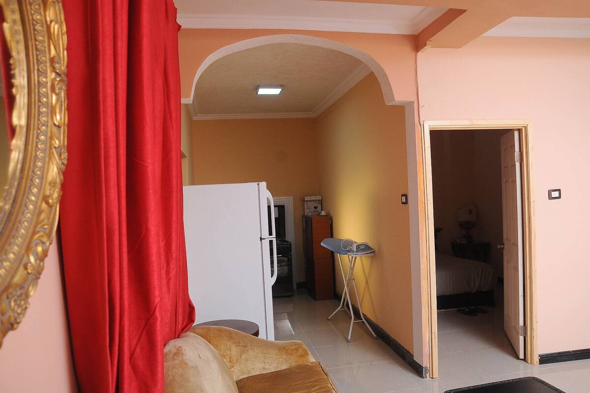 A room with a sofa, a dining table, a refrigerator, and a bed in the adjacent room.