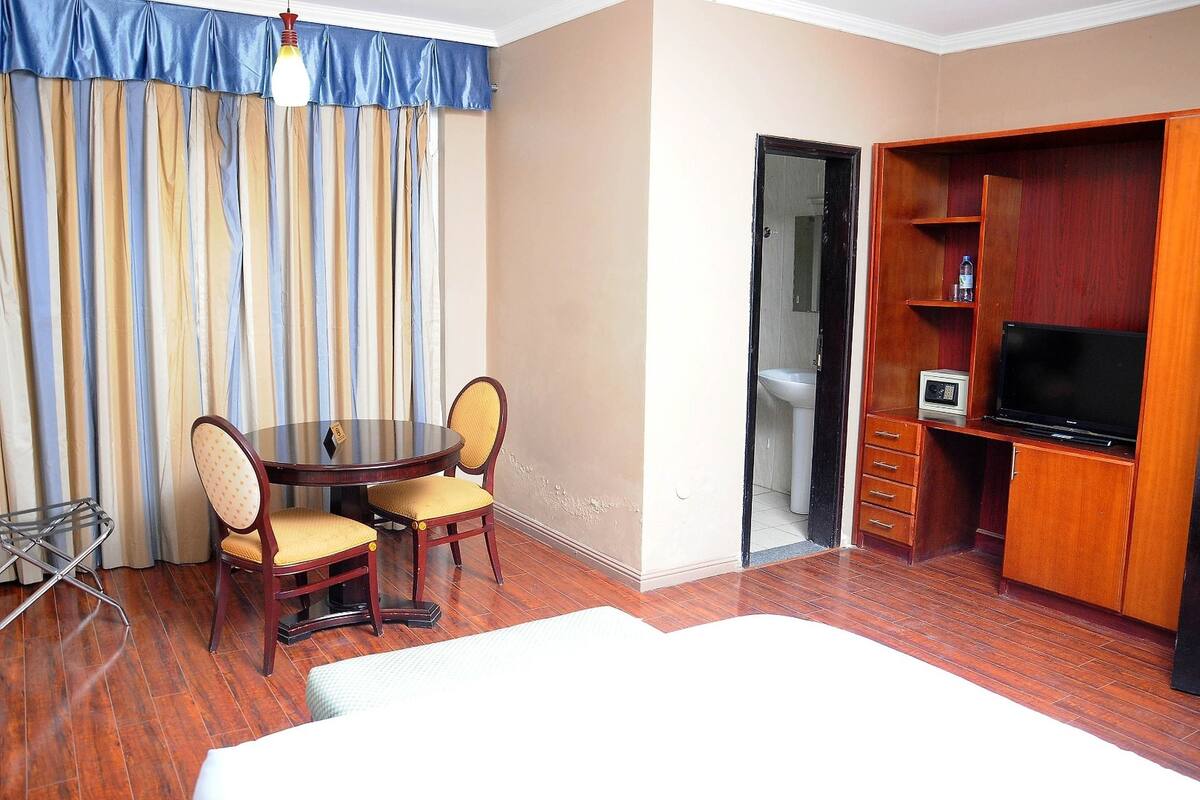 A hotel room with a wooden floor, a dining table with chairs, a TV, and a wardrobe.