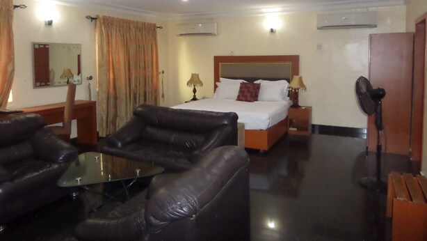 A hotel room with a bed, two bedside tables with lamps, a leather sofa, a glass coffee table, a standing fan, and a mirror on the wall. - Adna Hotel (Lagos)