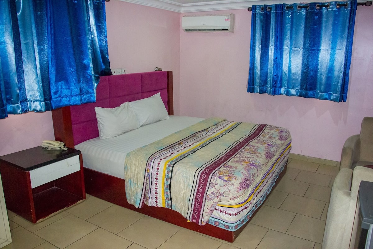 A bedroom with a bed, a nightstand, a chair, and a window with blue curtains.