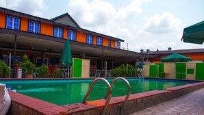 A swimming pool with metal railings, surrounded by a covered area with tables and chairs, and a building with orange and green walls in the background. - Esporta Suites (Lagos)