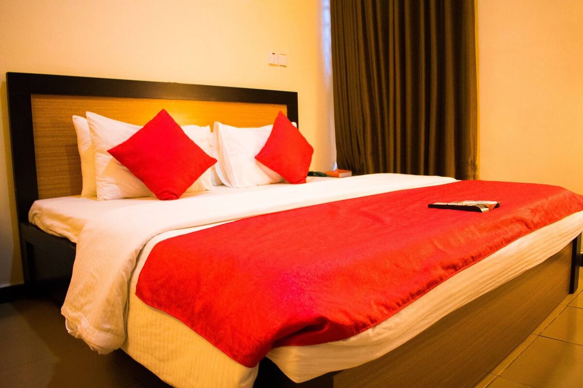 Una cama bien hecha con ropa blanca y roja, un control remoto sobre la cama y un cabecero oscuro.
