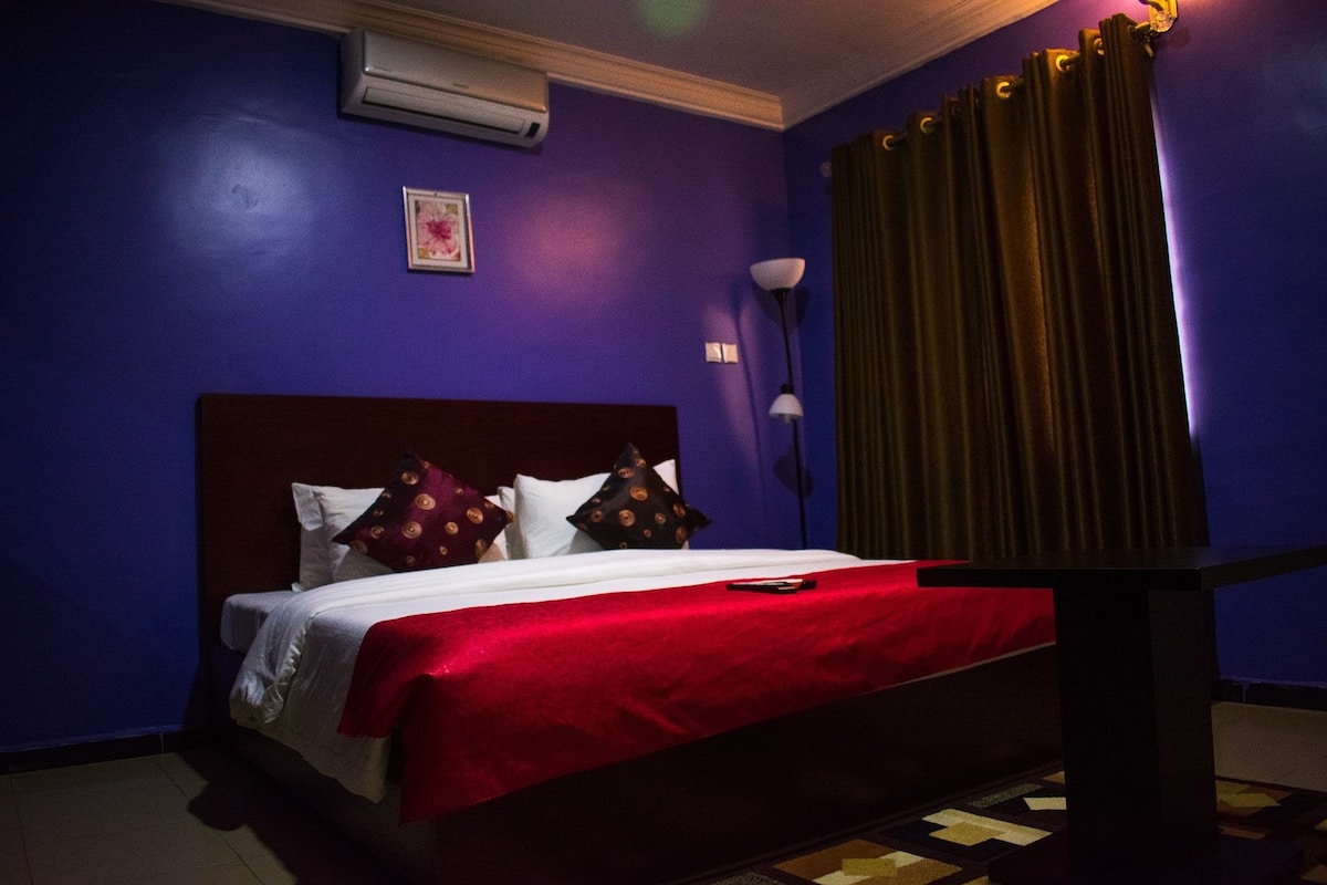 Habitación de hotel con colcha roja, cabecera de madera, una lámpara de noche y una ventana con cortinas.