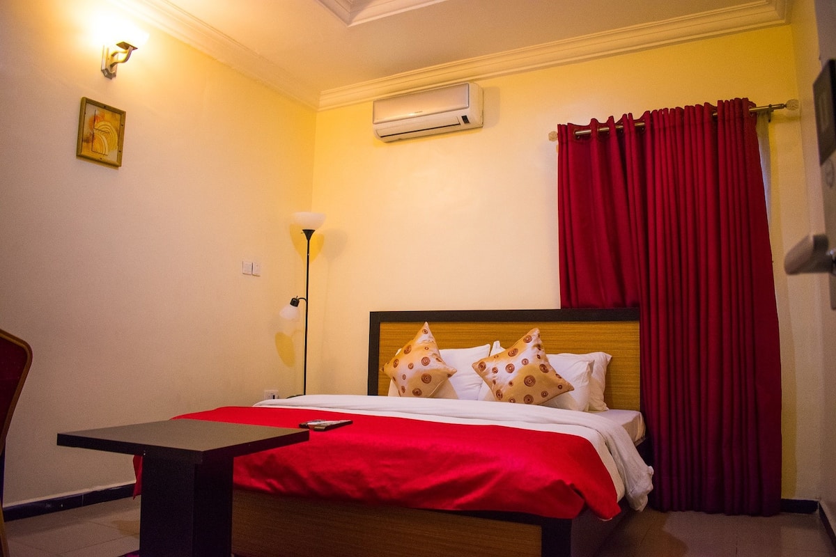 Habitación de hotel con una cama, una mesita de noche, una lámpara, aire acondicionado y una cortina roja.
