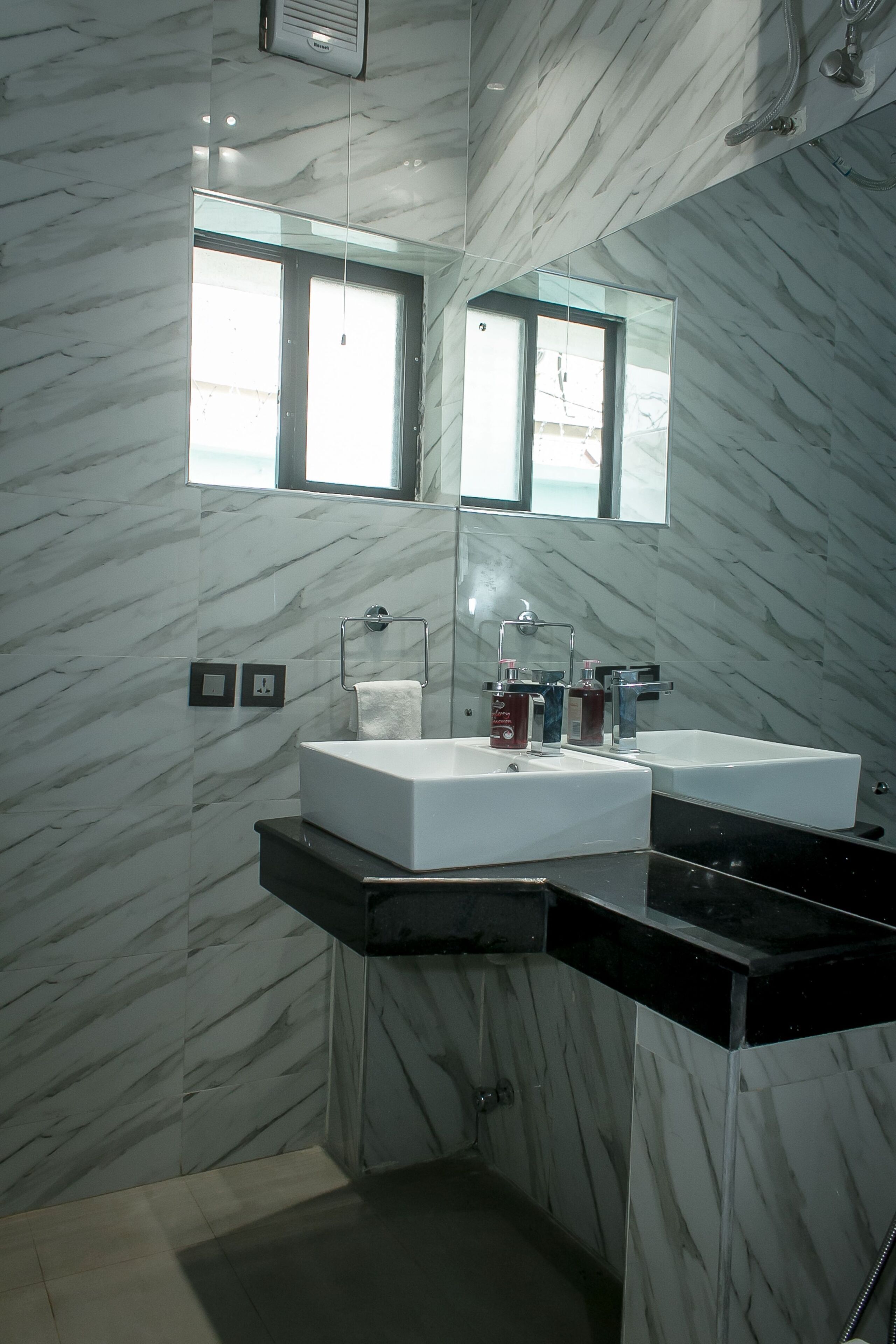 A modern bathroom with marble walls, a large mirror, two sinks, and a window providing natural light.
