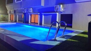 A swimming pool with a blue-lit deck, a covered area with a roof, and a building with multiple windows and air conditioning units. - Atlantic Hotel and Spa (Lagos)