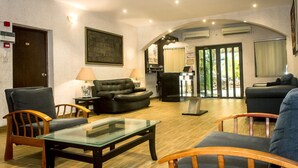 A spacious living room with a glass coffee table, two sofas, and a wooden armchair. There is a door, a lamp, and a painting on the wall. - Atlantic Hotel and Spa (Lagos)