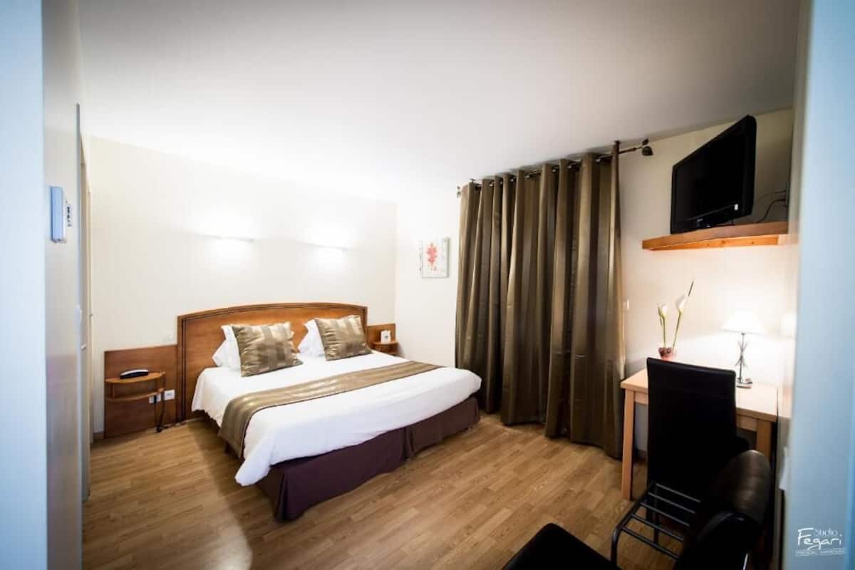 Une chambre d’hôtel avec un lit en bois, un bureau avec une chaise, un téléviseur fixé au-dessus du bureau et une fenêtre avec des rideaux.