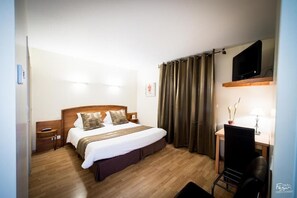 A hotel room with a wooden bed, a desk with a chair, a television mounted above the desk, and a window with curtains. - Hotel Le Lion D'or (Saint-Geniez-d'Olt-et-d'Aubrac)