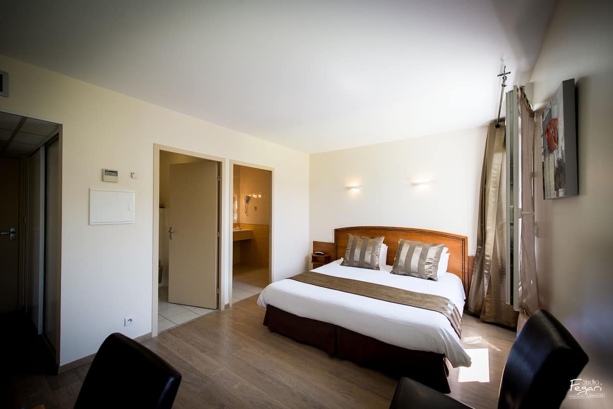 Une chambre d’hôtel avec un grand lit, une tête de lit en bois, une table de chevet et une chaise. Une porte donne accès à une salle de bain et à un couloir.