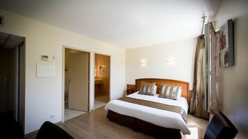 Une chambre d’hôtel avec un grand lit, une tête de lit en bois, une table de chevet et une chaise. Une porte donne accès à une salle de bain et à un couloir.