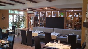 A restaurant with tables set for dining, a bar area with shelves of bottles, and a large window allowing natural light. - Hotel Le Lion D'or (Saint-Geniez-d'Olt-et-d'Aubrac)