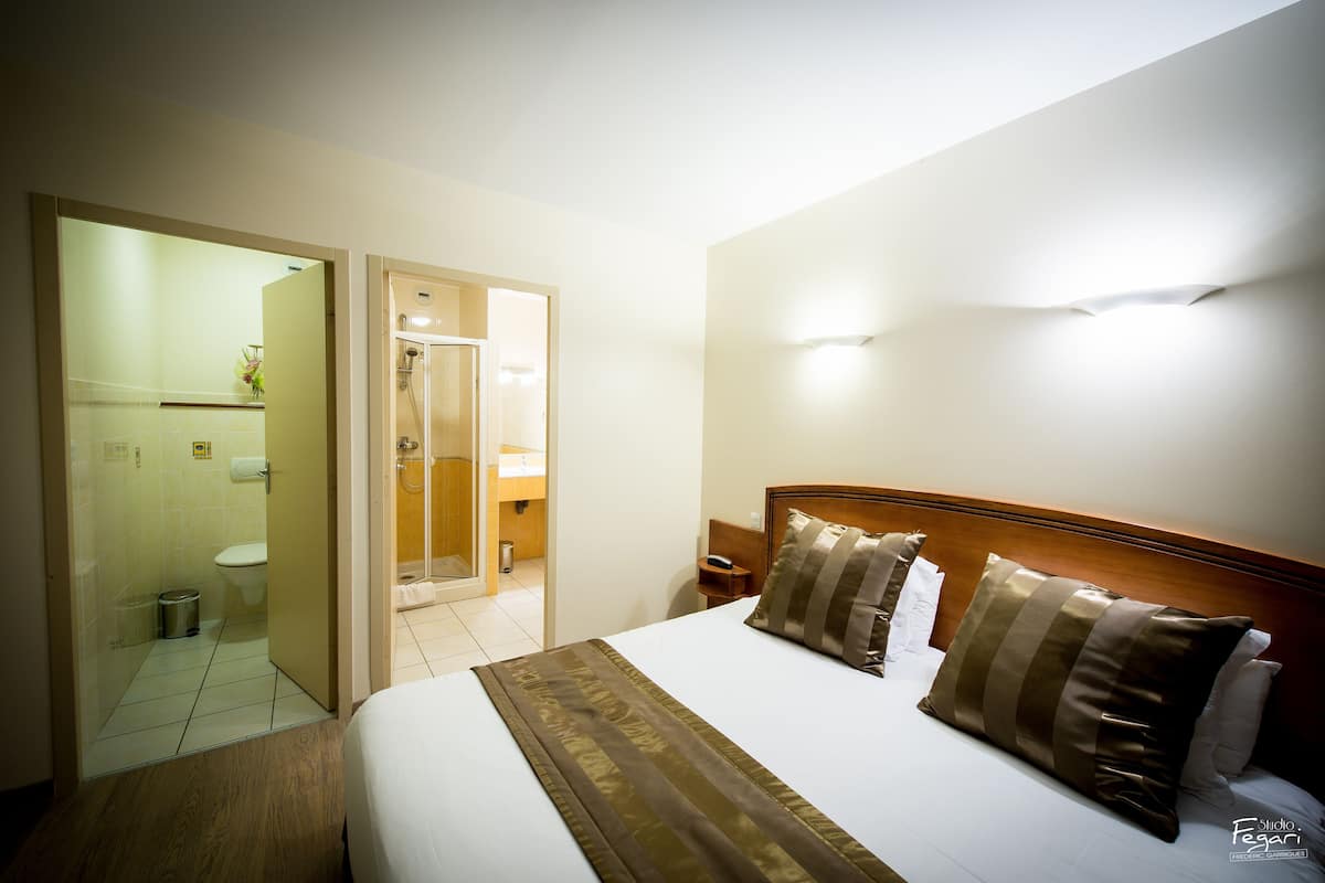Une chambre d’hôtel avec un lit, une table de chevet, une tête de lit en bois et une salle de bain visible à travers une porte ouverte.