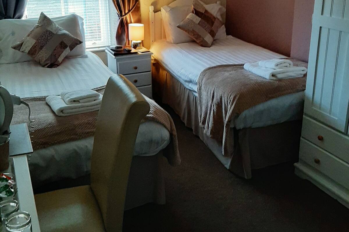 A hotel room with two beds, a desk, and a chair. There is a window with curtains, a lamp on the nightstand, and a framed picture on the wall.