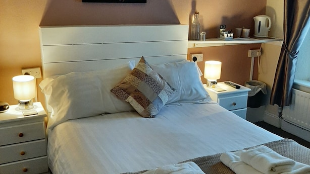 A neatly made bed with white linens, a decorative pillow, and folded towels on a woven mat. - Lyndhurst Guest House (Keswick)