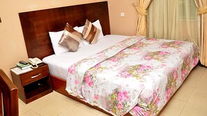 A neatly made bed with a floral bedspread, a wooden nightstand with a phone and books, and a window with curtains. - Golden Destiny Hotel Limited (Lagos)