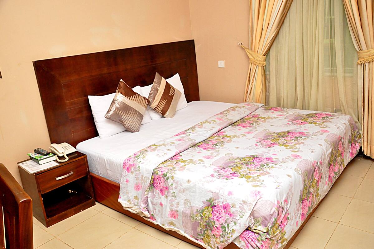 A neatly made bed with a floral bedspread, a wooden nightstand with a phone and books, and a window with curtains.