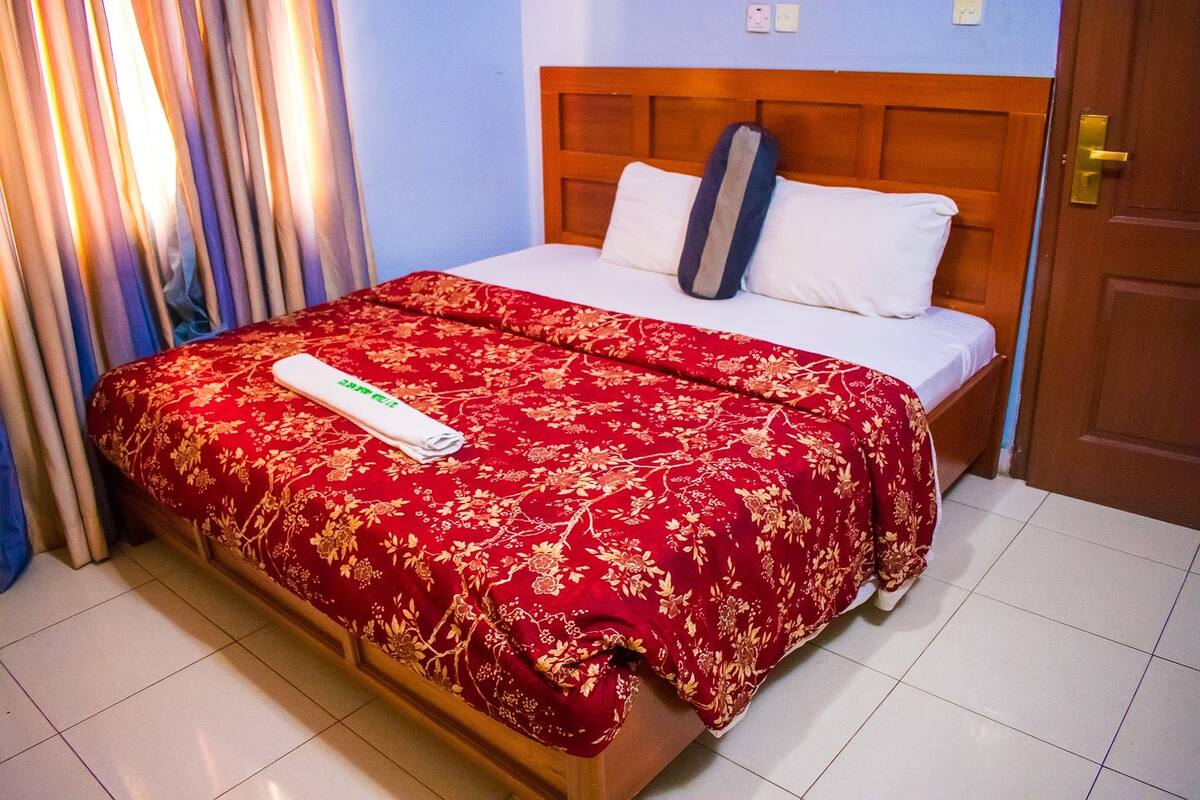 A neatly made bed with a red floral bedspread, a white pillow, and a grey pillow on a wooden headboard.