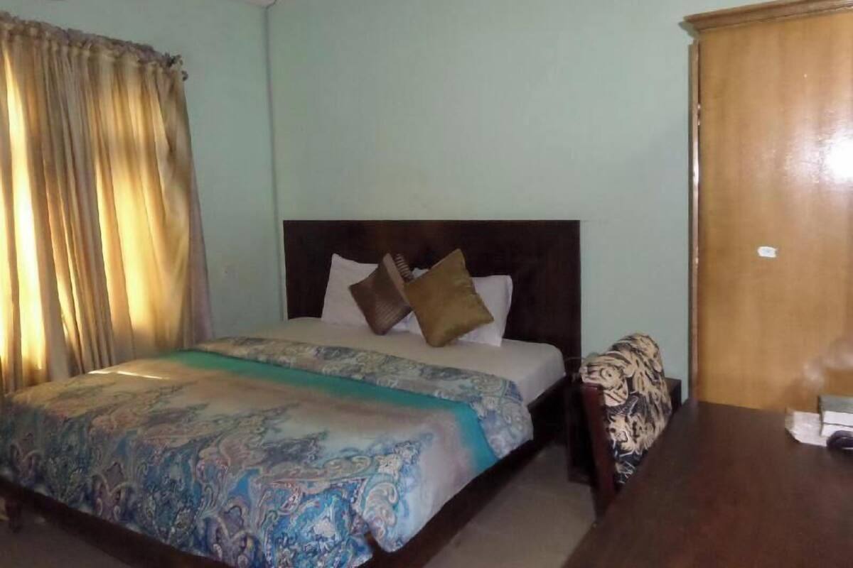 A bedroom with a bed, a wooden nightstand, a wardrobe, and a window with curtains.