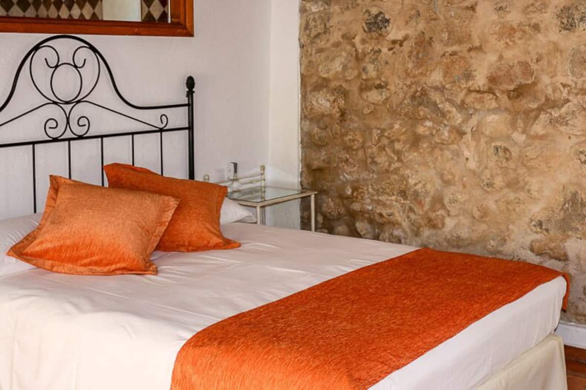 A bed with an orange blanket and pillows, a stone wall, and a metal headboard.