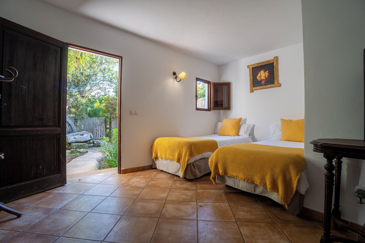 Un dormitorio con dos camas, un armario de madera, una ventana pequeña y una puerta que da a un área exterior.