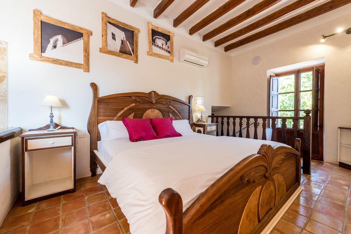 A bedroom with a wooden bed, a nightstand, a lamp, and a window with a view of greenery.