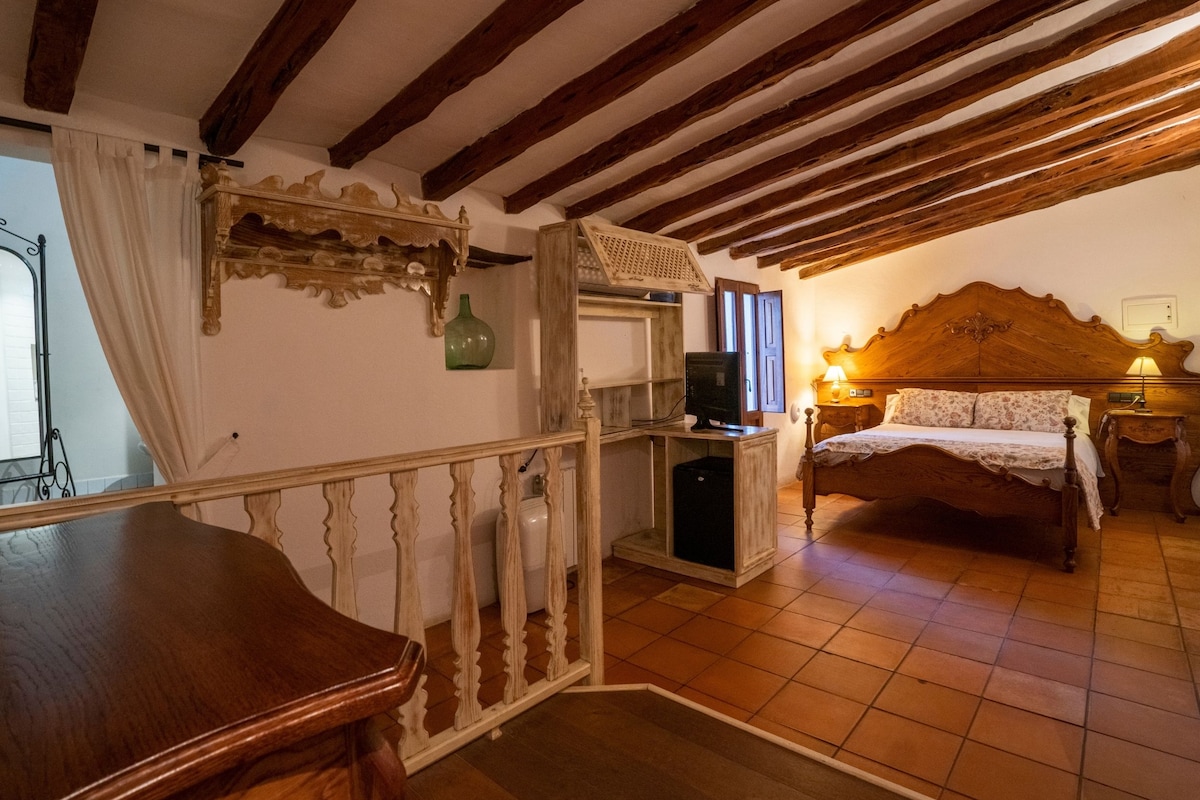 Un dormitorio con techo de madera, una cama con cabecera de madera, una mesita de noche con lámpara y un pequeño escritorio con computadora.