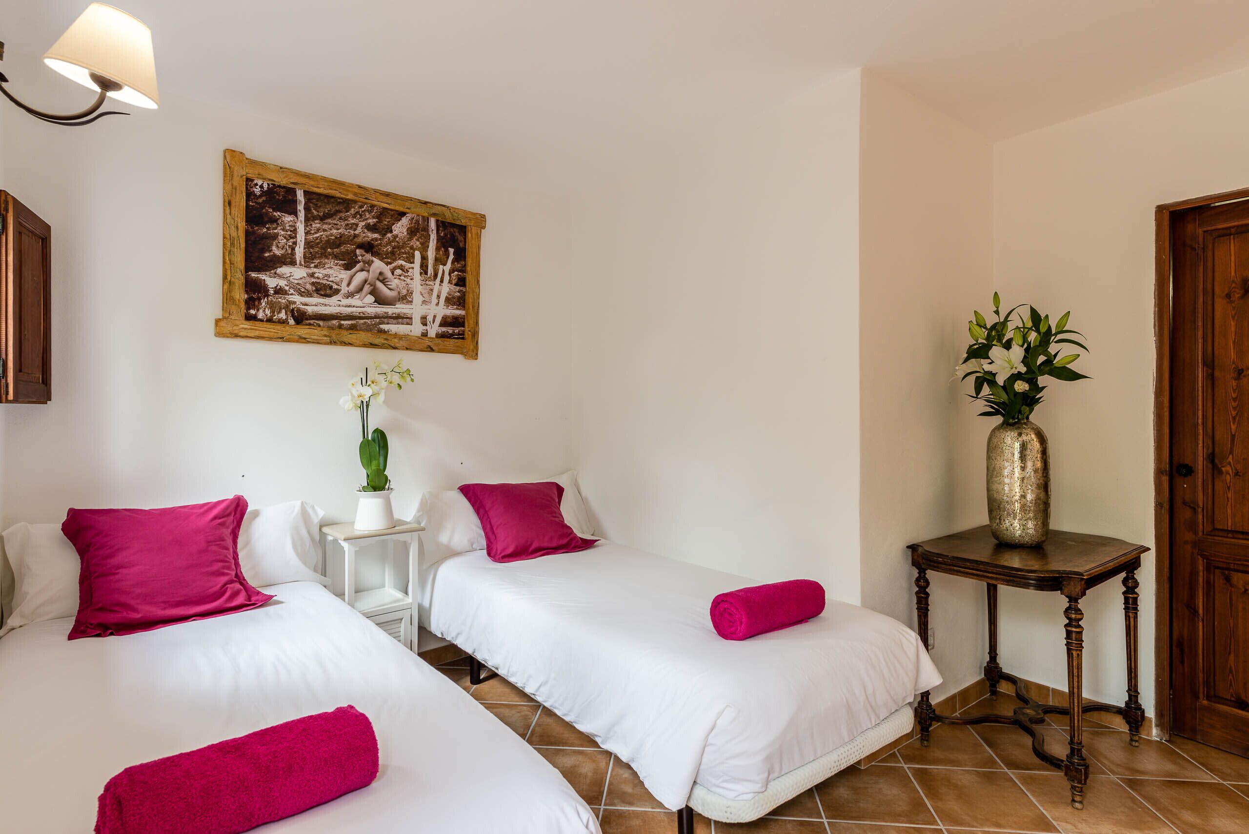 A hotel room with two beds, each with white bedding and pink pillows. There is a small table with a vase of flowers and a framed picture on the wall.