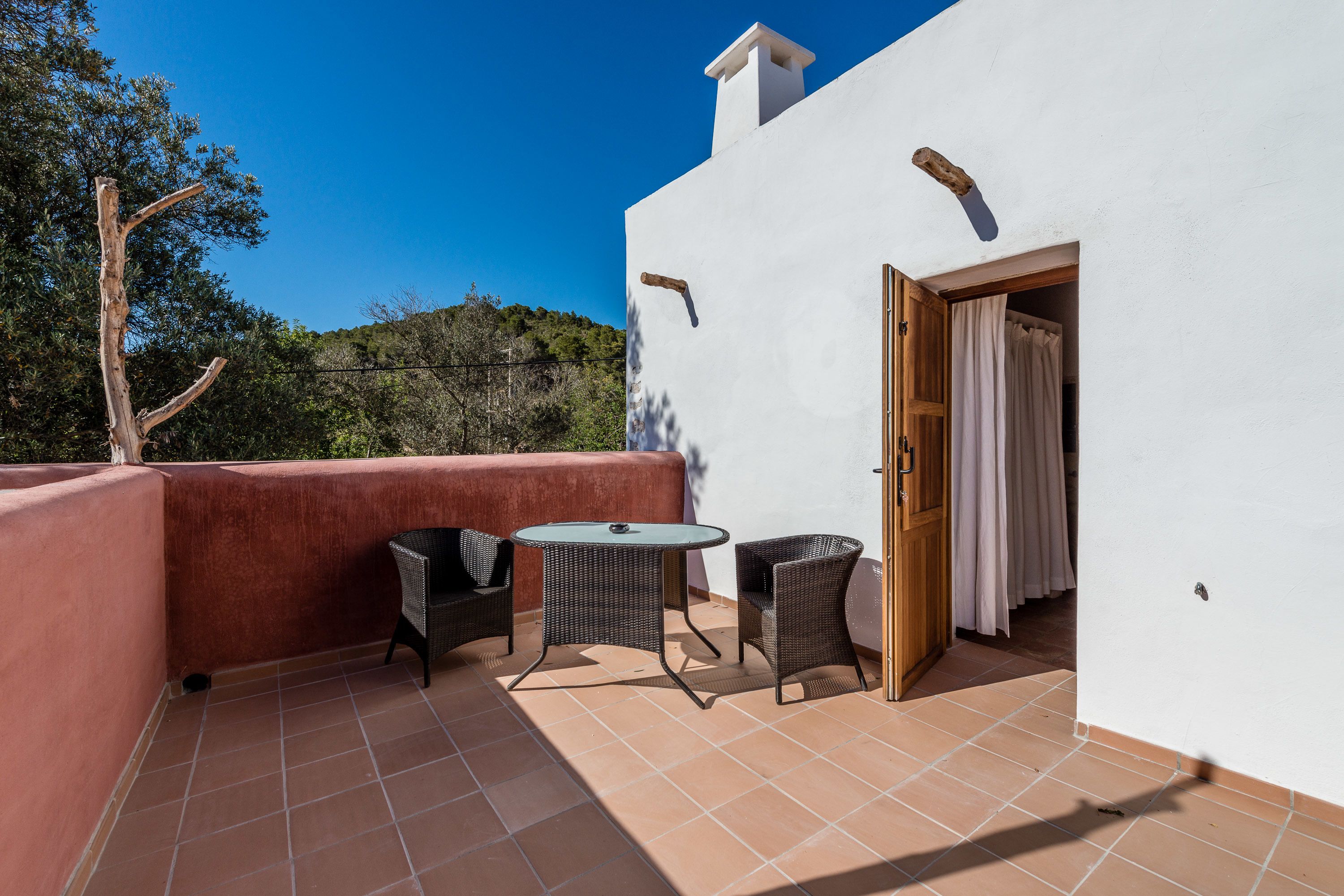 Un patio con dos sillas de mimbre y una mesa, un edificio blanco con un muro rojo, y una puerta con cortina.