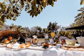 Un desayuno al aire libre con diversas opciones de comida y bebidas sobre una mesa, rodeado de abundante vegetación.