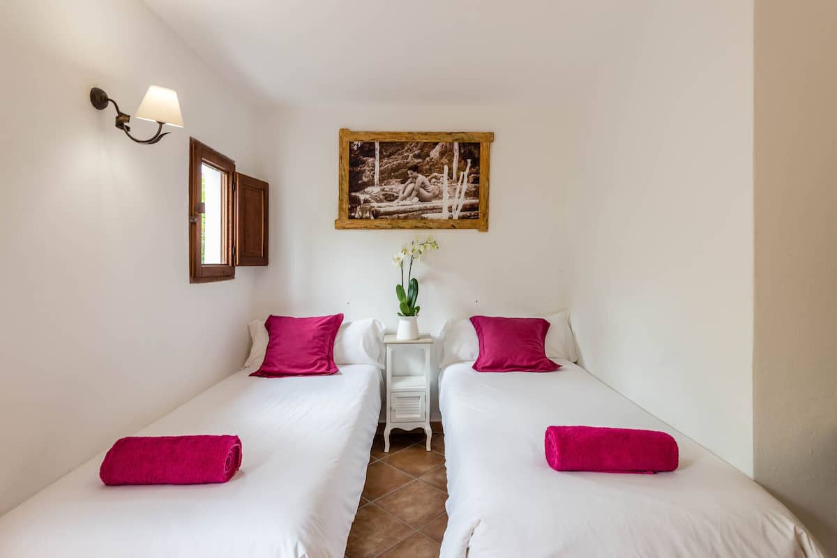 A hotel room with two single beds, each with a pink pillow and a rolled towel. There is a small white table with a plant between the beds. A framed picture hangs on the wall above the bed.