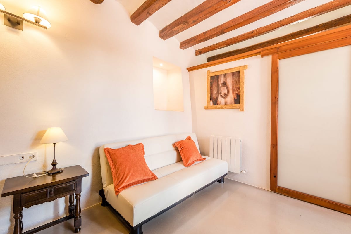 A room with a white sofa, two orange pillows, a wooden table with a lamp, a painting on the wall, and a radiator.
