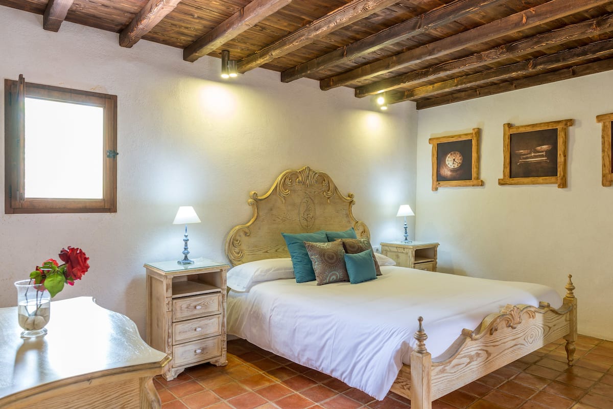 A bedroom with a wooden bed, two bedside tables with lamps, a nightstand, a window, and a wall clock.