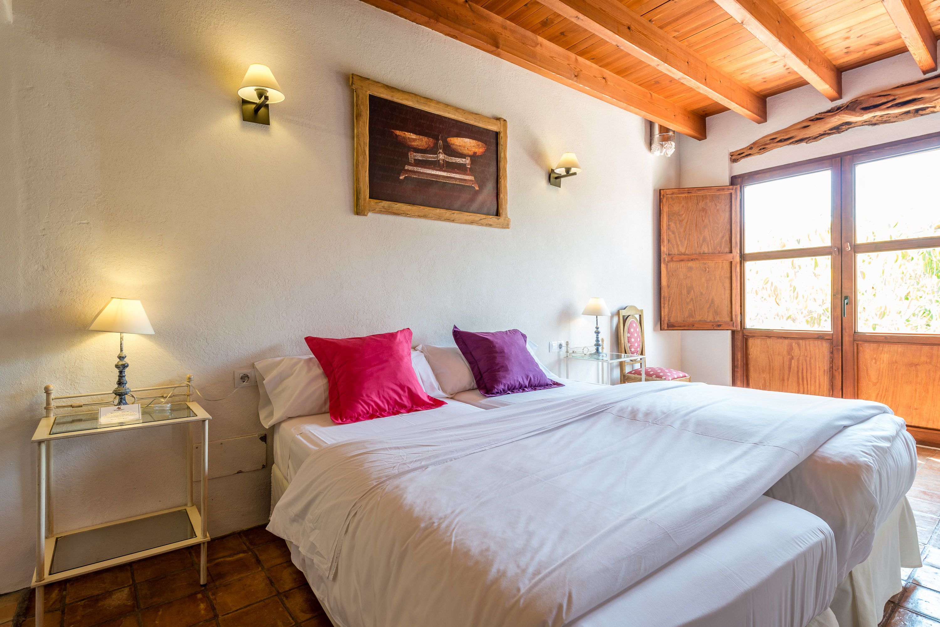 Un dormitorio con una cama grande, dos lámparas de noche, techo de madera y una ventana con vista a follaje.