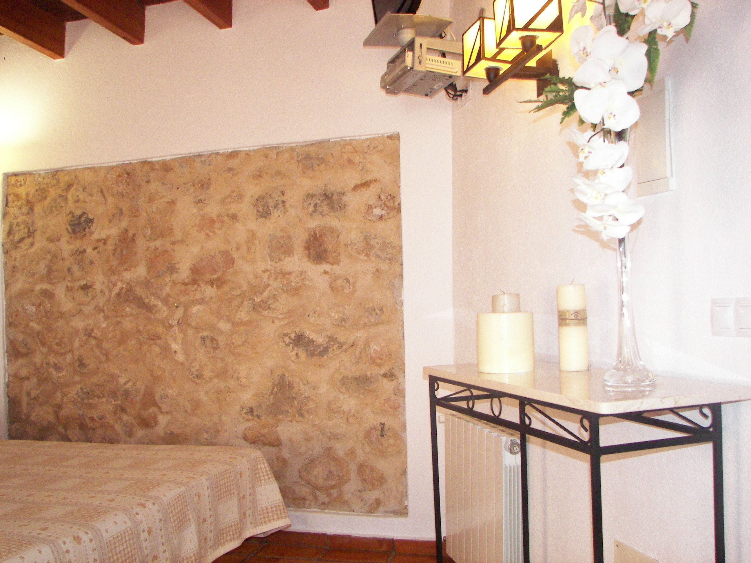 A bedroom with a stone headboard, a bed with a patterned bedspread, a small table with candles, and a vase with white flowers.
