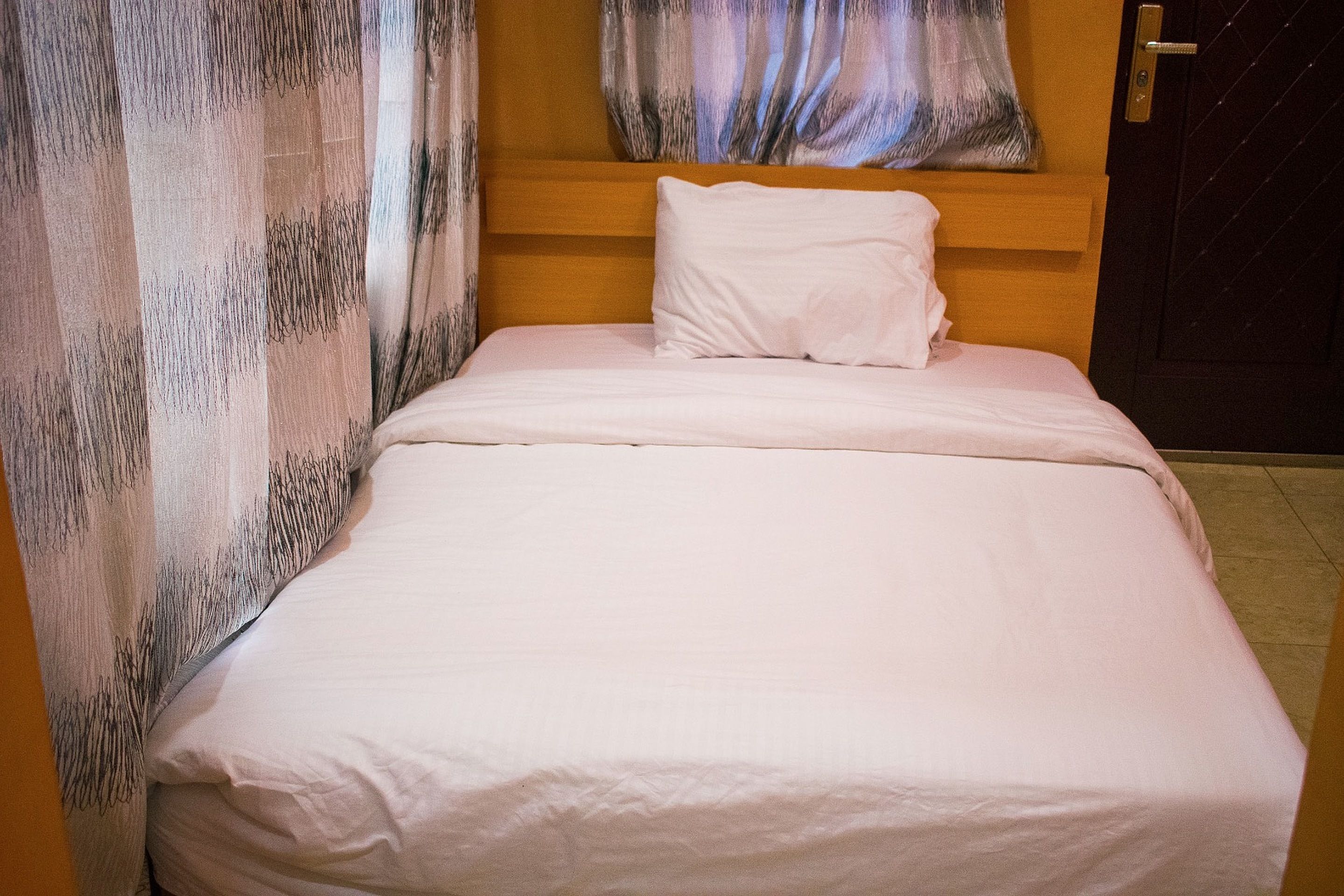 A neatly made bed with white bedding in a room with a wooden headboard and a patterned curtain.
