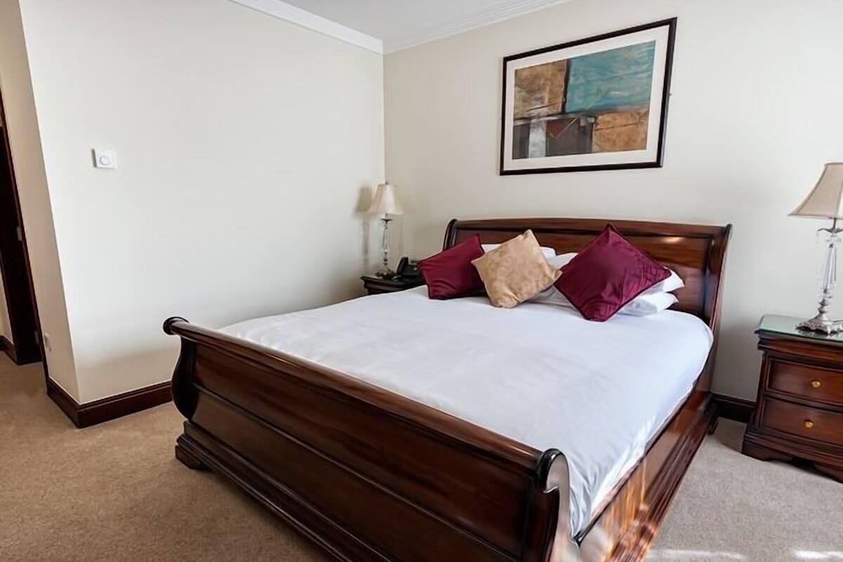 A neatly made bed with a wooden headboard, a bedside table with a lamp, and a painting on the wall.