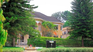 Une grande maison avec un toit en tuiles, entourée d’une végétation luxuriante et agrémentée d’une petite fontaine.