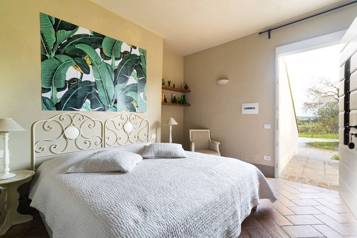 A bedroom with a large bed, a decorative headboard, a painting of tropical leaves, a small table with a lamp, and a view of the outdoors through an open door.