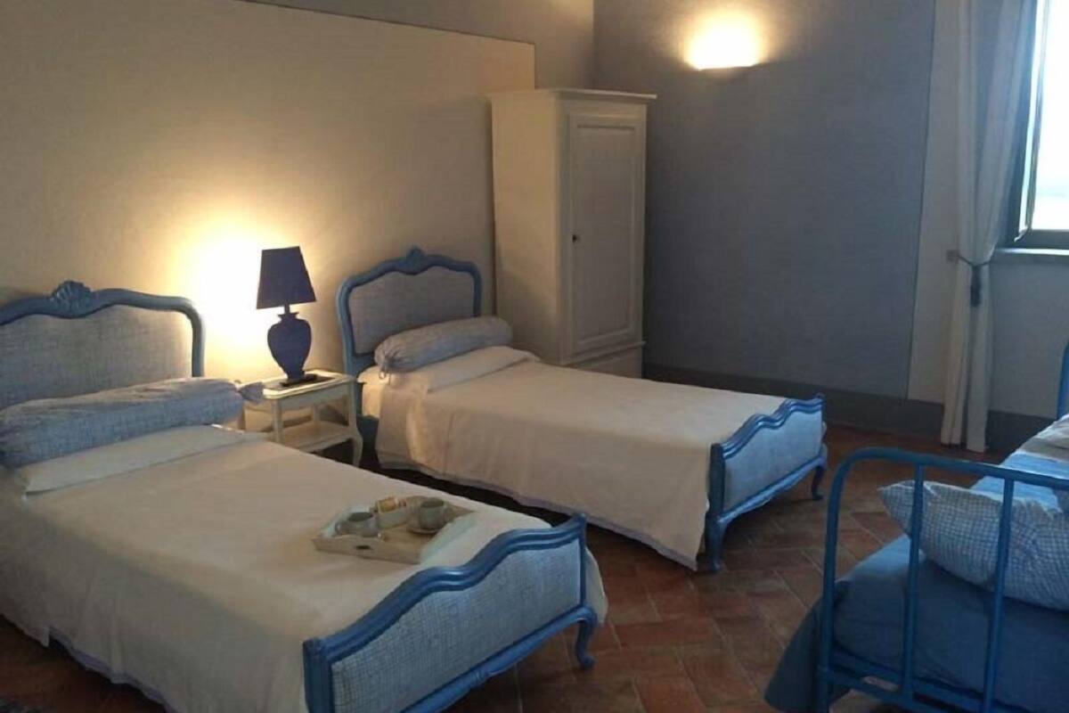 A room with two beds, a nightstand with a lamp, a wardrobe, and a window with curtains.