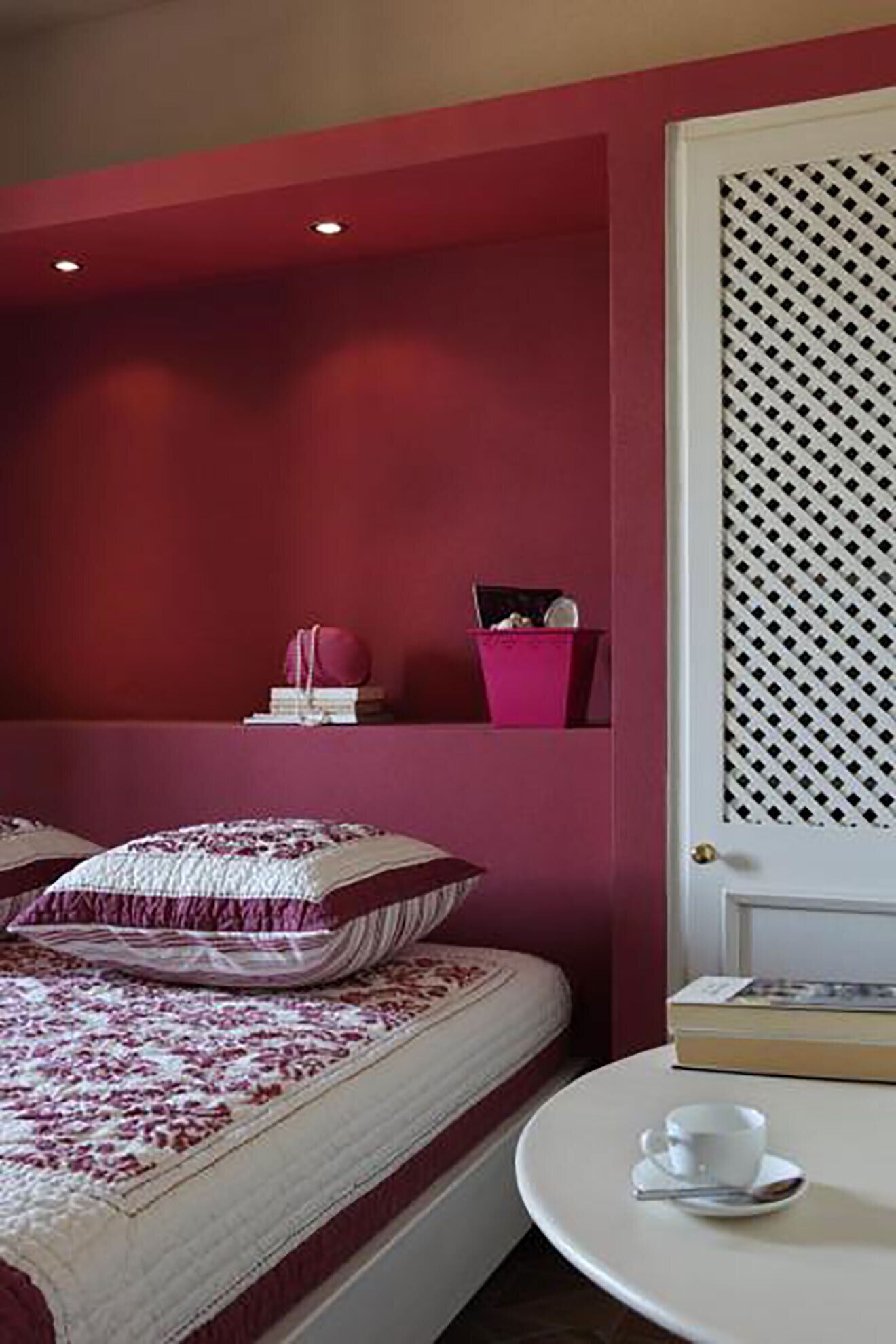 Camera da letto con un letto, cuscini, un comodino con tazza e piattino, e una piccola mensola con libri e un oggetto rosa.