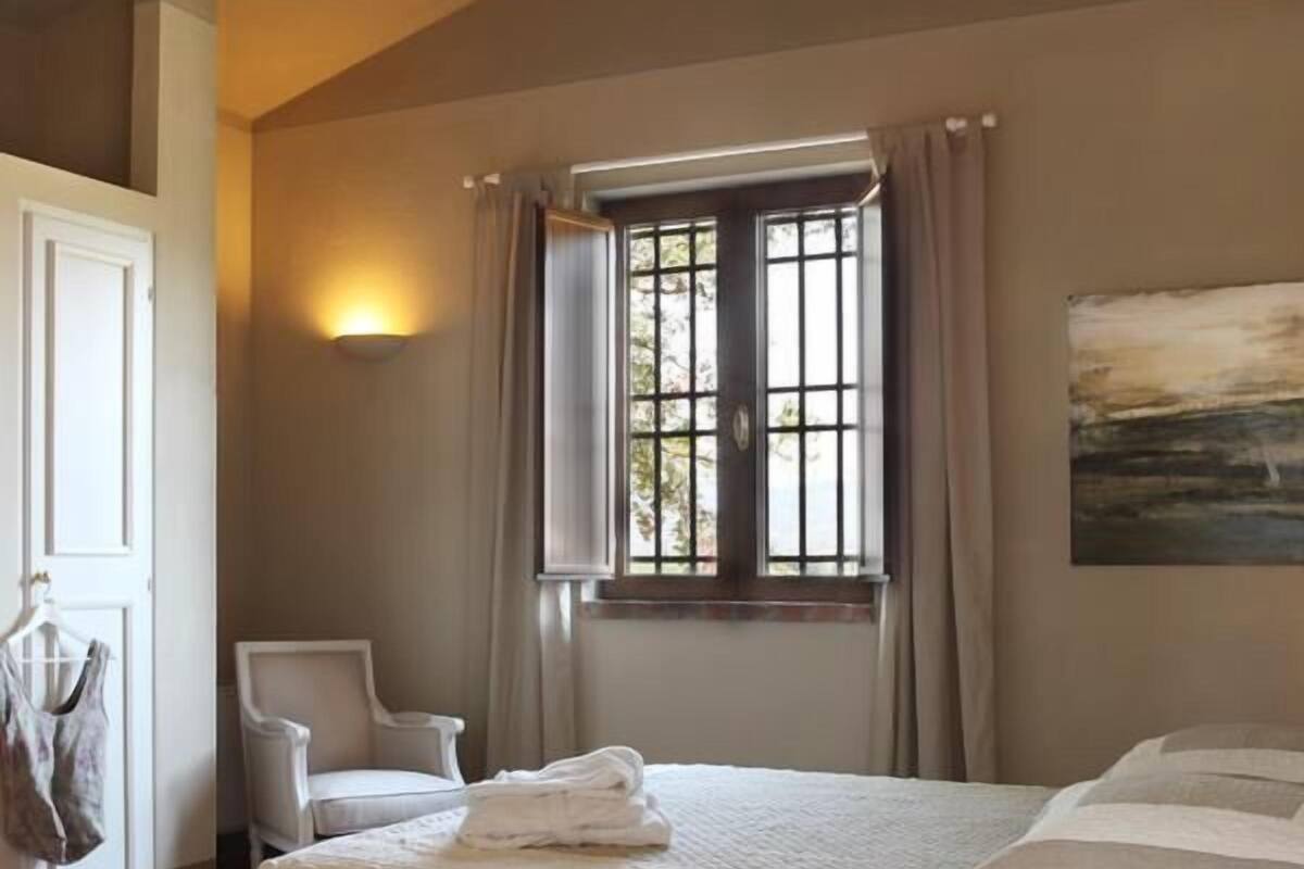 A bedroom with a bed, a chair, a table, a window with curtains, and a painting on the wall.