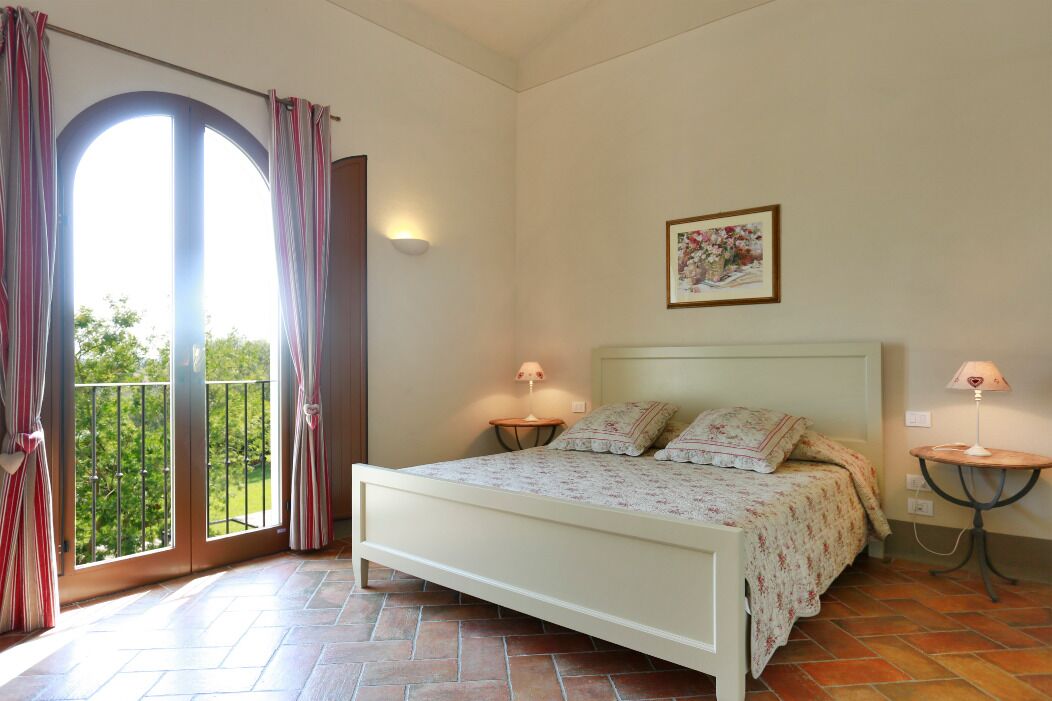 A bedroom with a bed, two bedside tables, a wall-mounted lamp, a picture, and a balcony with curtains.