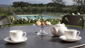 A table set with white cups and saucers, a clear glass cake stand with pastries, and a teapot, with a pool and greenery in the background. - Agriturismo Cantagrillo (Vinci)